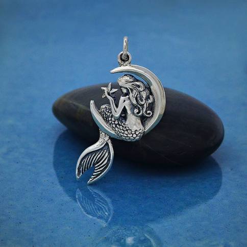 The Mermaid on the Moon pendant by Nina Designs features a sterling silver mermaid with shimmery scales sitting on a crescent moon, holding a star, set against black stone and a blue background.