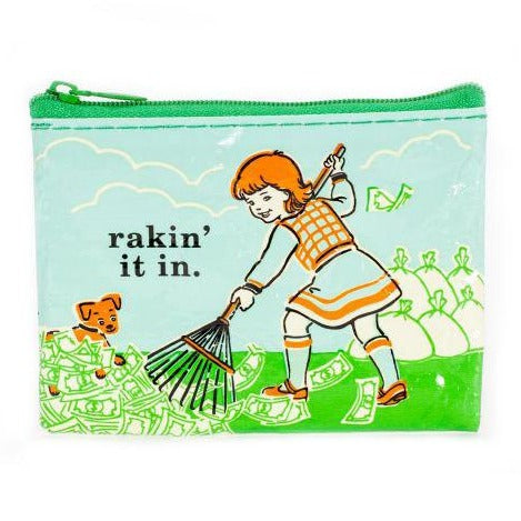 A girl in an orange dress rakes up money as a dog watches. Beside her, the text “rakin’ it in.” appears, with recycled-material money bags behind. This is featured on the Blue Q Raking It In Coin Purse.