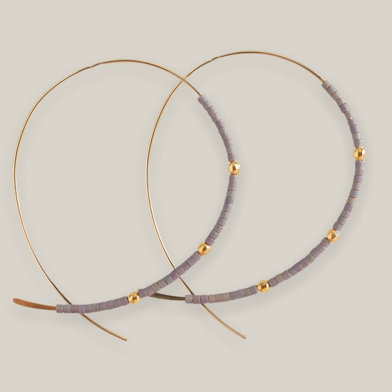The Confetti Earrings, Purple by Lenny & Eva are large gold hoops adorned with small lavender Miyuki beads and spaced gold plated beads, shown on a light beige background.