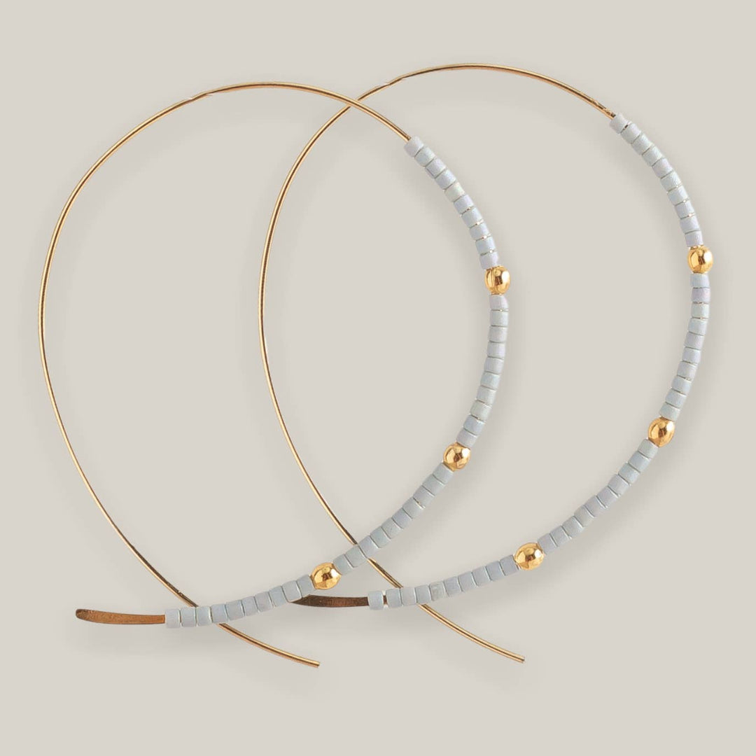 The Confetti Earrings, Grey by Lenny & Eva feature large gold-plated hoops with pale gray Miyuki beads and spaced gold accents, shown against a neutral beige background.