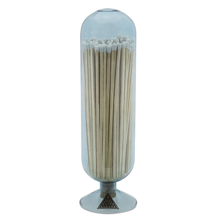 The Skeem Fireplace Match Cloche Sky is a tall, hand-blown glass container on a silver metal base, filled with long wooden matches featuring white tips.