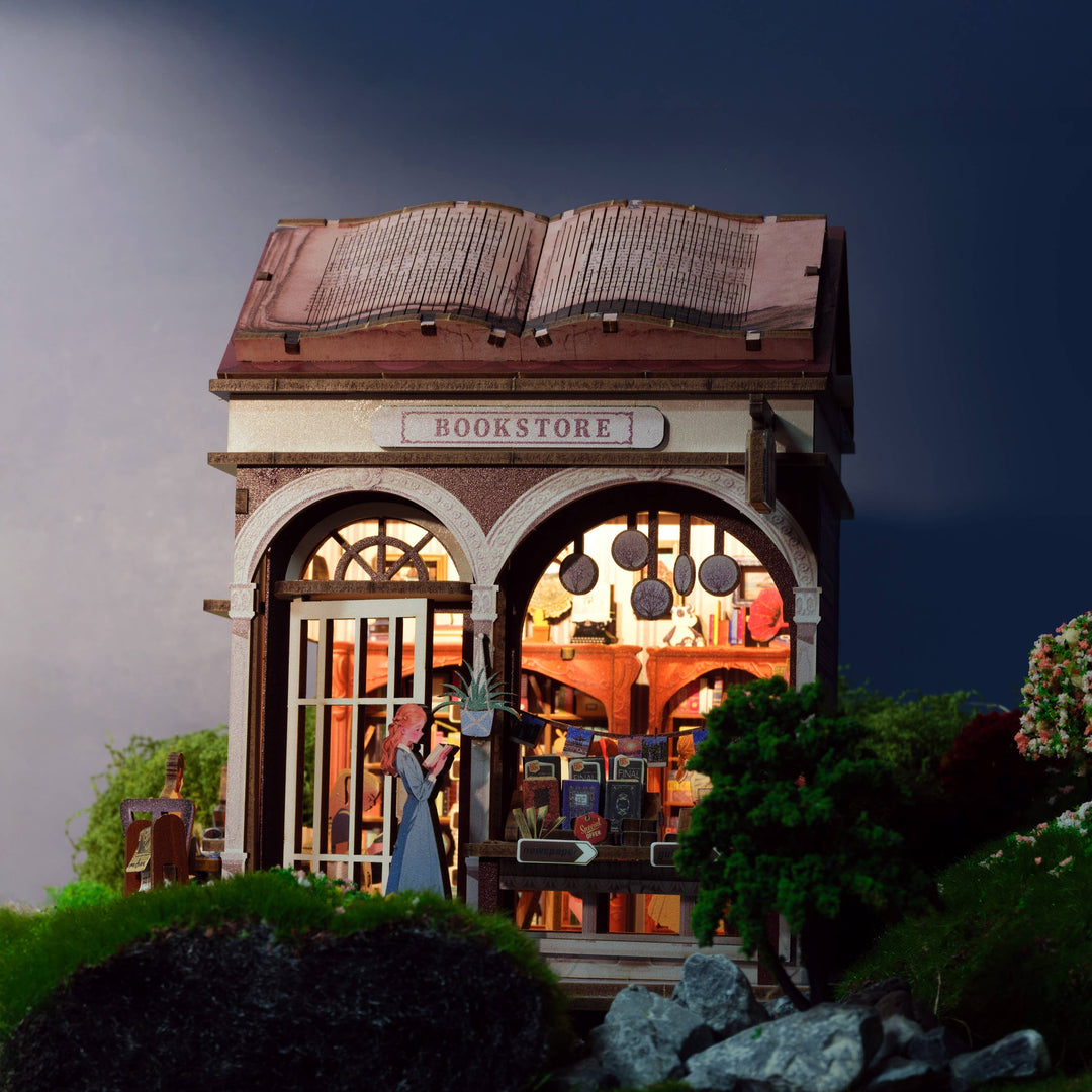 The Hands Craft DIY Miniature House Kit: Bookstore features a tiny shop with an open book roof, arched windows, a reading figure, and cottagecore details like trees, grass, and rocks—all set against a striking dark background.