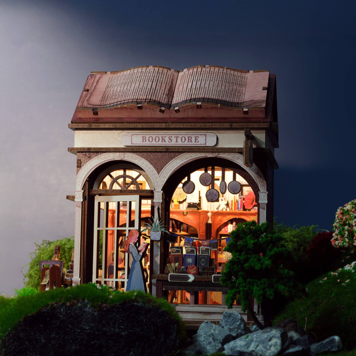 The Hands Craft DIY Miniature House Kit: Bookstore features a tiny shop with an open book roof, arched windows, a reading figure, and cottagecore details like trees, grass, and rocks—all set against a striking dark background.
