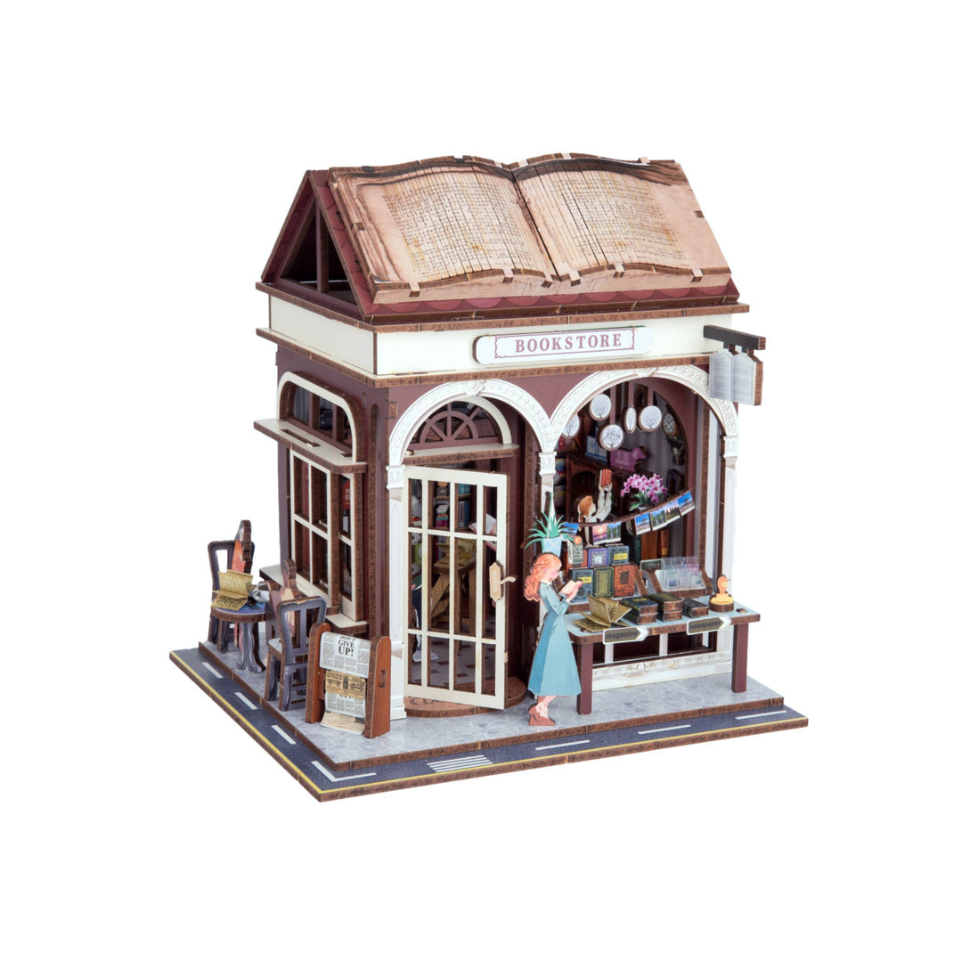 The Hands Craft DIY Miniature House Kit: Bookstore features a cozy shop with arched windows, outdoor books, a table and chairs, and an apron-clad figure arranging items—ideal for cottagecore fans.