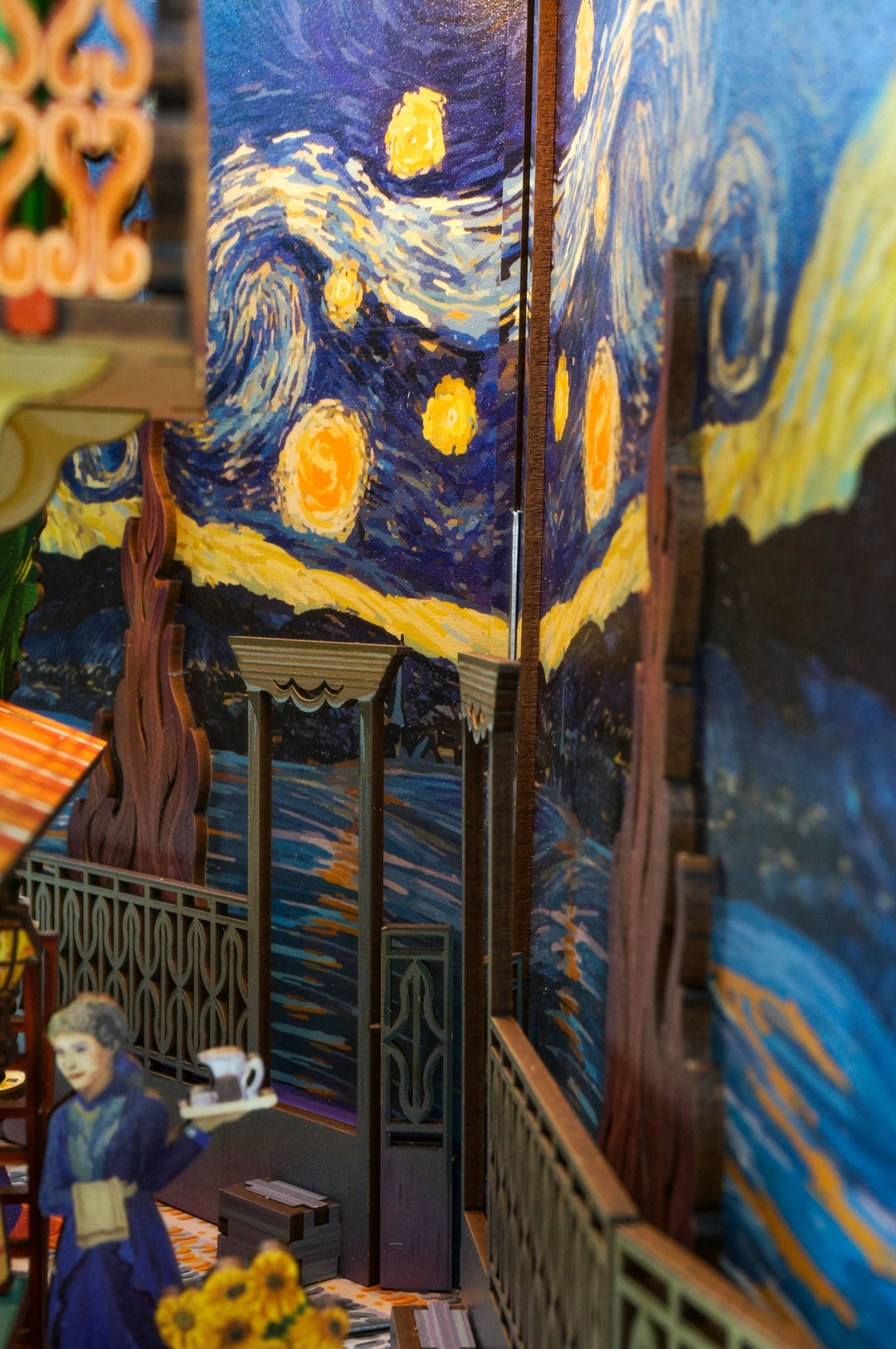 A close-up of Hands Craft’s DIY Book Nook Kit: Vincent's World shows a café scene with a paper figure and a Van Gogh-inspired "Starry Night" bookshelf insert, where yellow stars swirl in the night sky above.