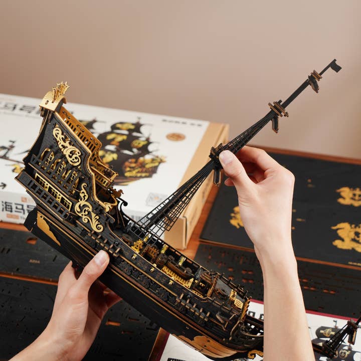 A person assembles the Hands Craft DIY The Seahorse Barque Ship, a detailed black and gold model, holding it above a table with assembly sheets and packaging in the background.