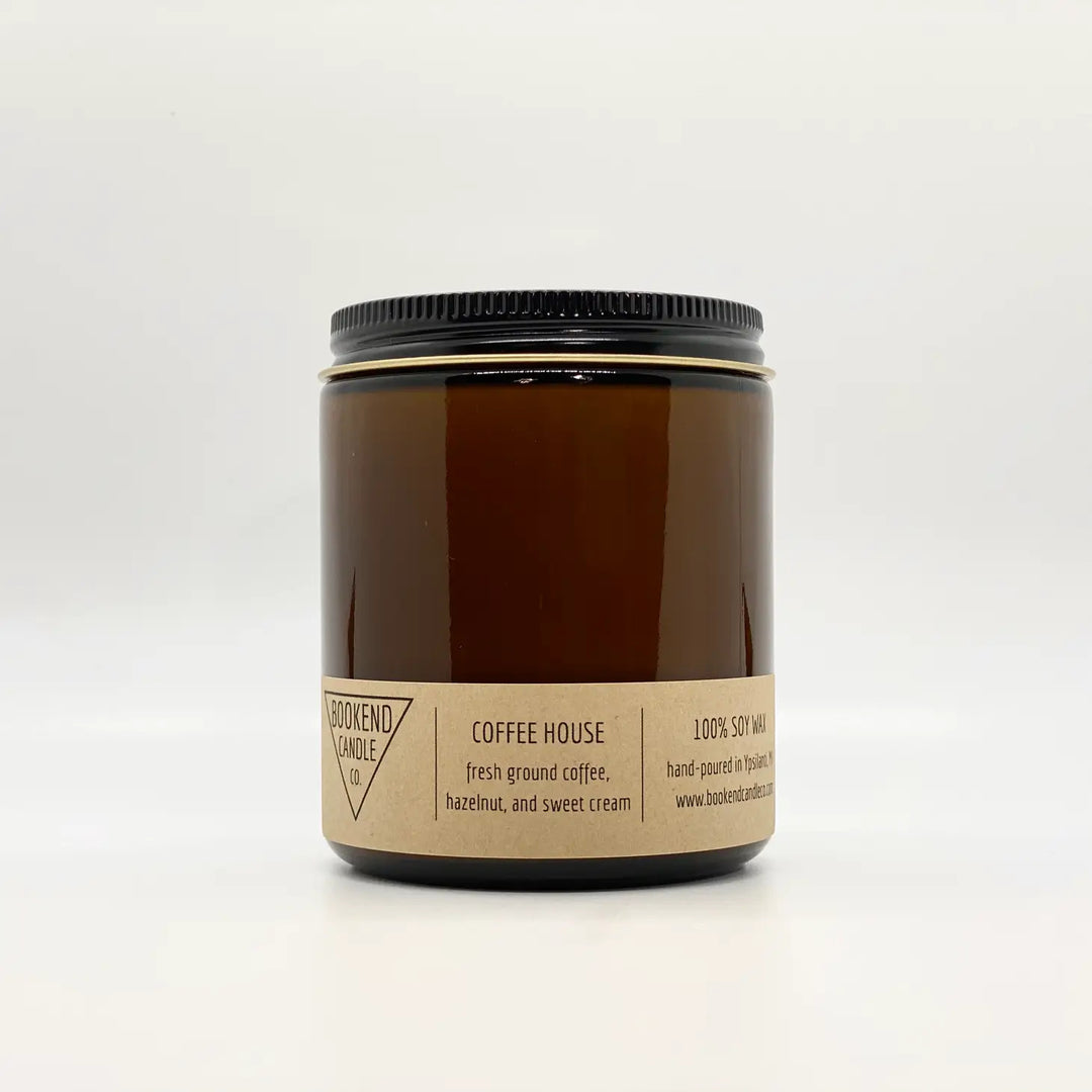 An amber glass jar with a black lid contains the "Coffee House Soy Candle 8oz" by Bookend Candle. The label reads "100% soy wax," "hand-poured in Franklin, MA," and lists the scent notes as "fresh ground coffee, hazelnut, and sweet cream." This candle features cotton wicks and phthalate-free fragrances.