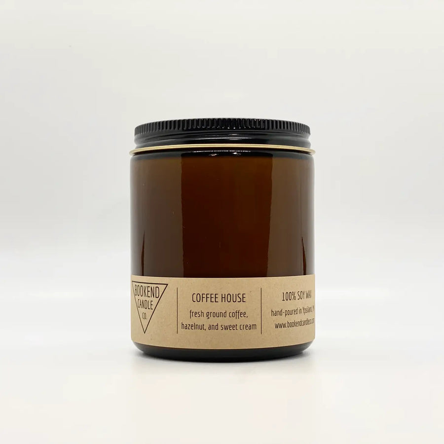 An amber glass jar with a black lid contains the "Coffee House Soy Candle 8oz" by Bookend Candle. The label reads "100% soy wax," "hand-poured in Franklin, MA," and lists the scent notes as "fresh ground coffee, hazelnut, and sweet cream." This candle features cotton wicks and phthalate-free fragrances.