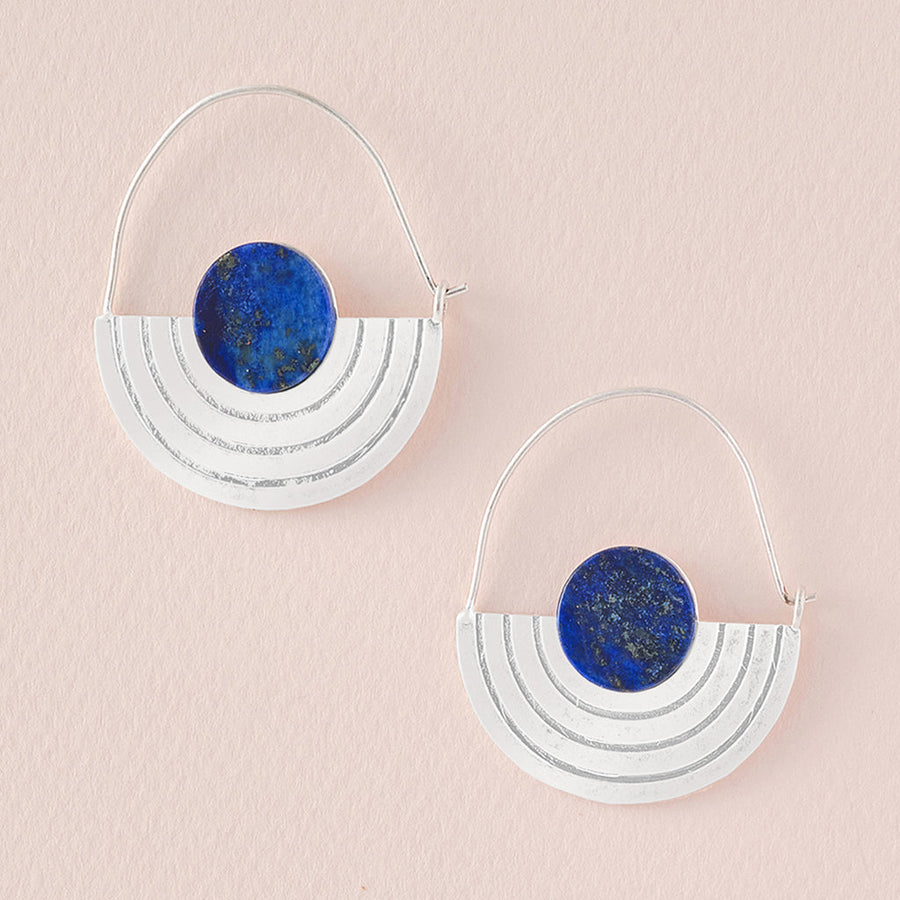 Two Stone Orbit Earrings with a modern geometric design from Scout Jewelry lie on a light pink surface. Each silver, hypoallergenic earring features a deep blue lapis circular stone at the top, set above three arched silver bands that form a semi-circular shape. These minimalistic earrings give a striking and elegant look.