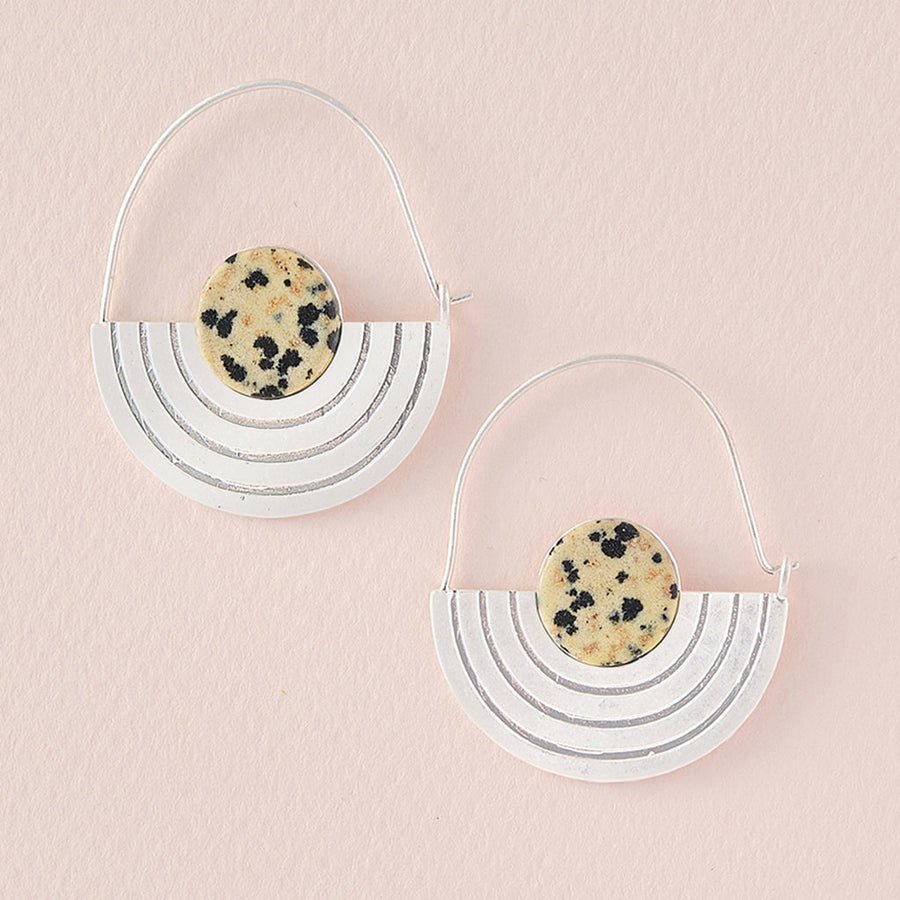 The Stone Orbit Earring Dalmatian Jasper Silver by Scout Jewelry is a pair of minimalistic earrings set against a soft pink background. Each earring showcases a semi-circle silver frame adorned with engraved lines and topped with a round dalmatian jasper gemstone, mimicking the distinctive dalmatian pattern. This ensures they are not only stylish but also hypoallergenic, making them perfect for any occasion.