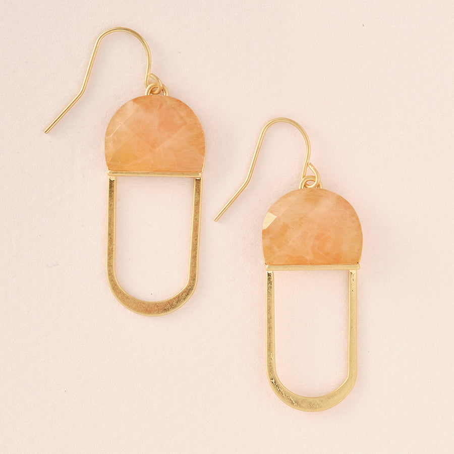 The Modern Stone Earring Sunstone Gold by Scout Jewelry showcases a geometric design, featuring 14K gold-dipped dangle earrings with a semi-circular sunstone set atop a rectangular frame. These hypoallergenic earrings have hook fastenings and are presented against a light beige backdrop.