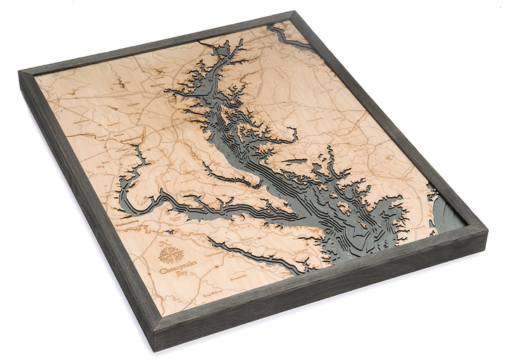 The WoodChart Chesapeake Bay 24.5" x 31" Grey Frame features a framed wooden map of the bay and its region, with detailed carved inlets, rivers, and land elevations in natural wood tones.