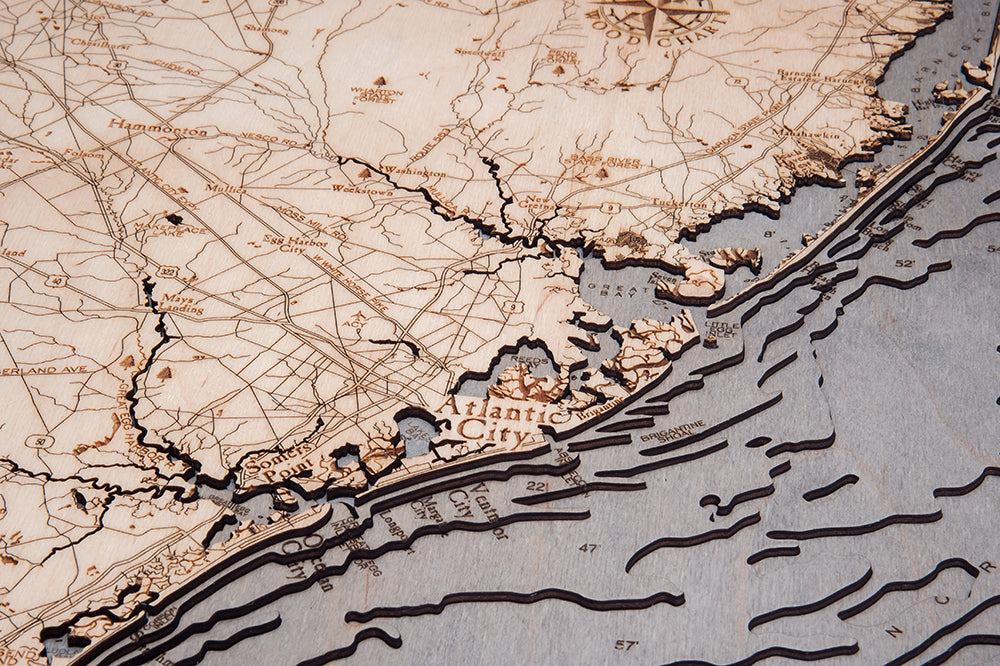 The WoodChart NJ Full Shore 24.5 x 31 Grey Frame is a detailed carved wooden map of Atlantic City’s coastline and inland, featuring labeled roads, cities, and topographic lines depicting land and ocean features.
