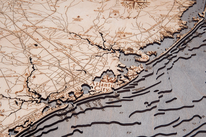 The WoodChart NJ Full Shore 24.5 x 31 Grey Frame is a detailed carved wooden map of Atlantic City’s coastline and inland, featuring labeled roads, cities, and topographic lines depicting land and ocean features.