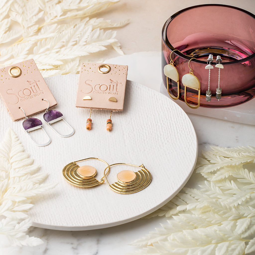 A display of Scout Jewelry's Stone Orbit Earrings in Sunstone Gold on a white plate and a pink glass dish showcases 14K gold-dipped accents, pearls, and vibrant stones like sunstone. Elegant white fern leaves decorate the marble surface surrounding this stylish arrangement.