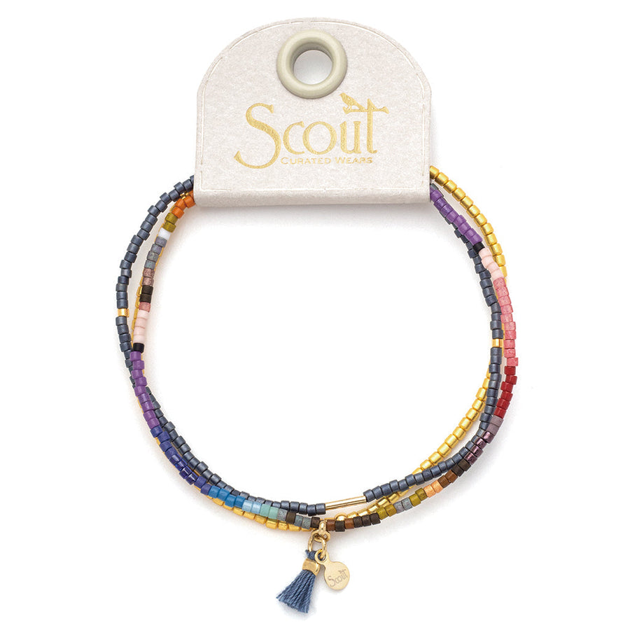 The Dark Multi Gold Miyuki Bracelet Trio from Scout Jewelry features a vibrant array of colorful Miyuki Delica beads and is adorned with a gold charm, complemented by a tiny blue tassel. It is beautifully presented on a branded card with a rounded top, making it perfect for layering elegance.