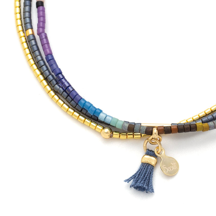 Introducing the Dark Multi Gold Miyuki Bracelet Trio by Scout Jewelry, featuring an array of vibrant blue, purple, and gold Miyuki Delica beads. This striking bracelet set is elegantly highlighted with a 14K gold heart charm and a dainty blue tassel.