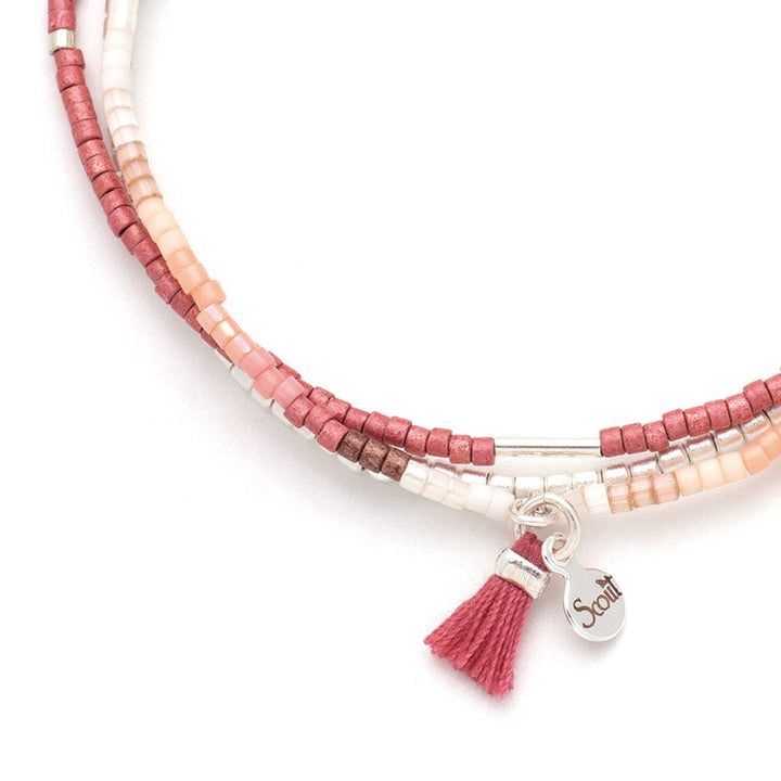 The Blush Multi Silver Miyuki Bracelet Trio by Scout Jewelry is showcased, crafted with pink, red, and white Miyuki Delica beads on a stretch cord. It features elegant silver accents with a pink tassel charm and a silver tag engraved with "Scout," ideal for stacking bracelet enthusiasts.