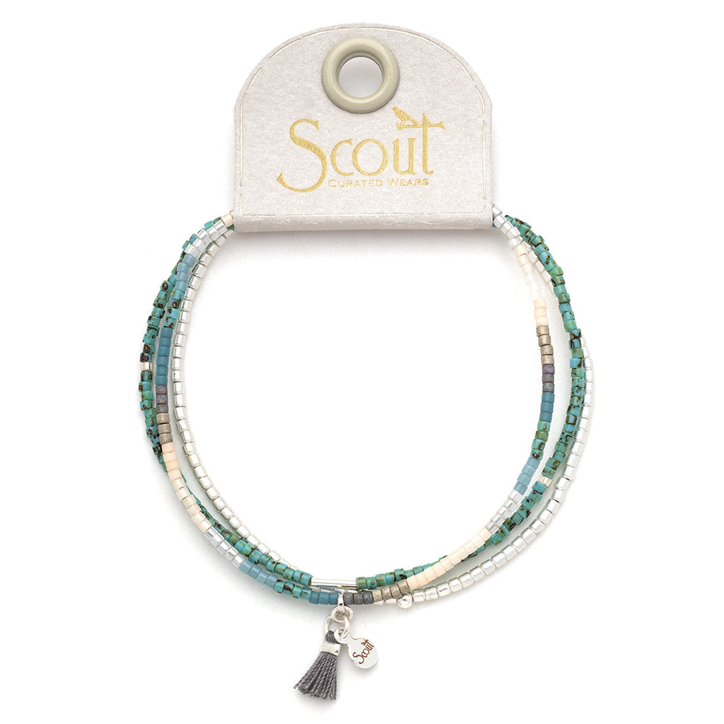 The Turquoise Multi Silver Miyuki Bracelet Trio from Scout Jewelry is displayed on a card backing. This beaded stacking bracelet features small Miyuki Delica beads in shades of turquoise, blue, white, and metallic silver, and includes a small tassel charm and a round pendant charm.