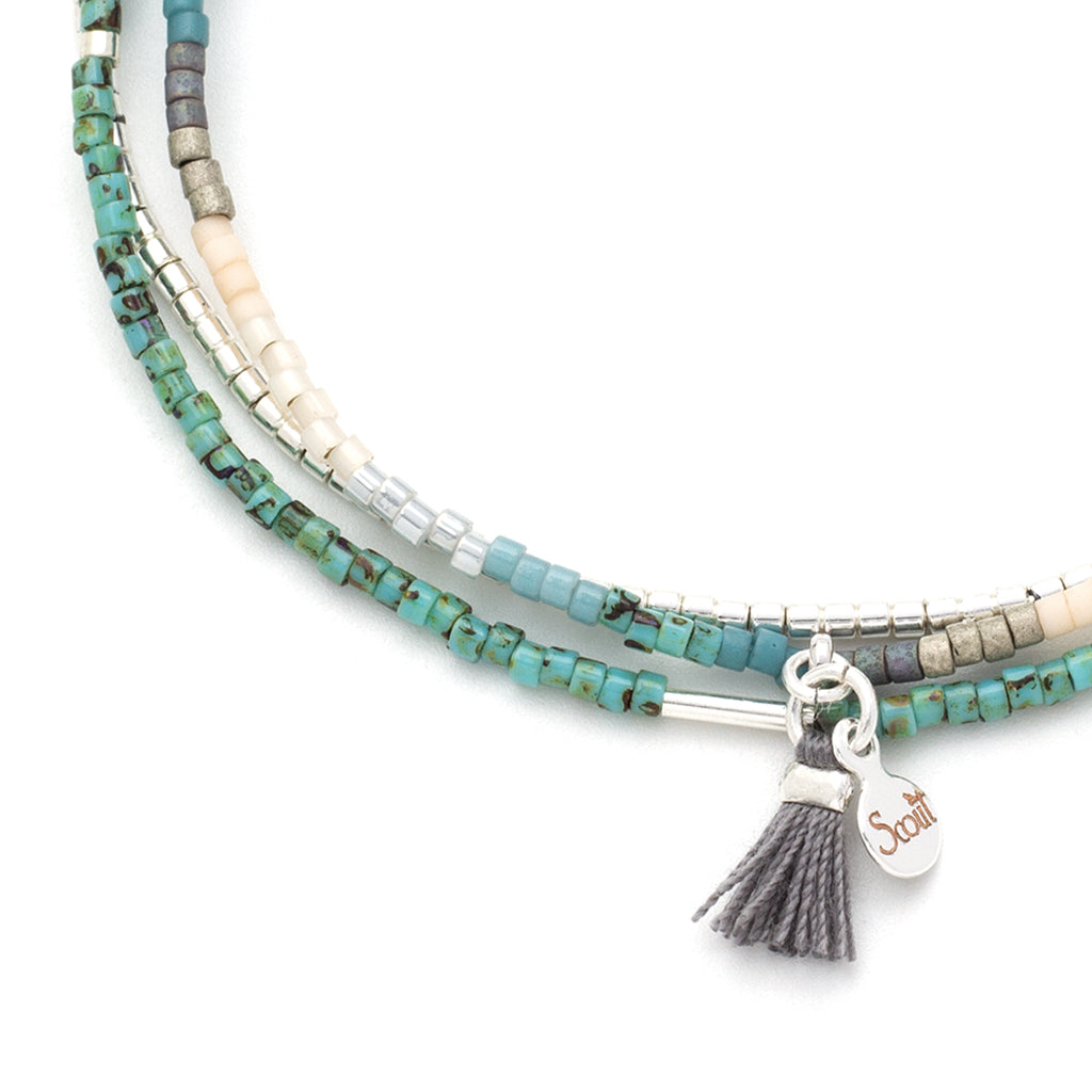 A close-up of the Turquoise Multi Silver Miyuki Bracelet Trio from Scout Jewelry reveals a multistrand design with small turquoise, silver, and beige Miyuki Delica beads. The bracelet features a small silver tag engraved with "Scout," alongside a grey tassel charm, making it an excellent choice for layering with other stacking bracelets for a stylish, layered look.