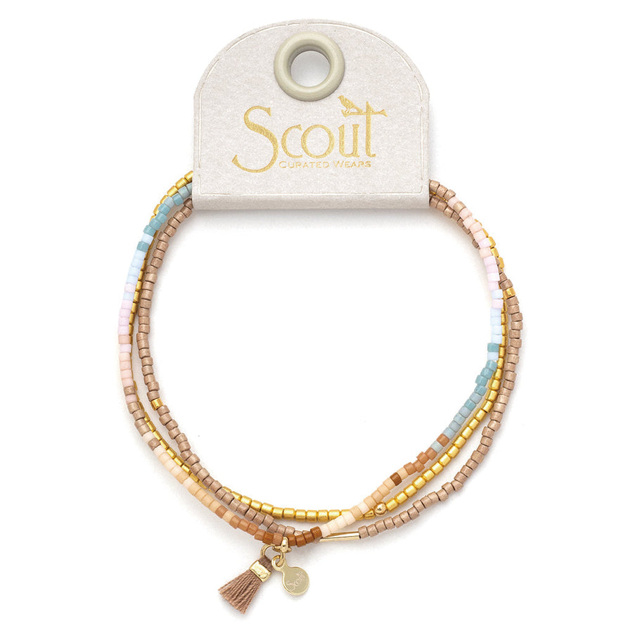 The Desert Multi Gold Miyuki Bracelet Trio by Scout Jewelry is displayed on a beige card and features multiple strands of small, colorful Miyuki Delica beads in shades of pink, blue, gold, and beige. A small gold tassel charm adorns the bottom of the card. Perfect for stacking bracelets, the brand name "Scout" is printed on the card.