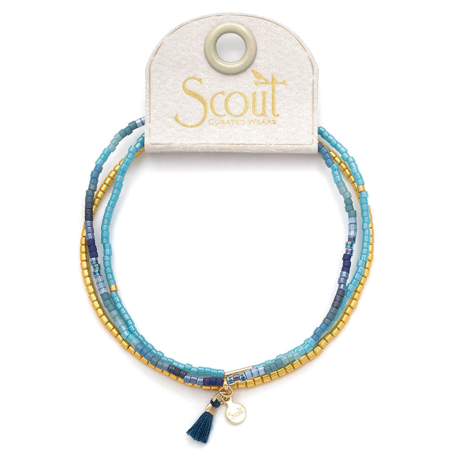 A beaded bracelet set titled "Cobalt Multi Gold Miyuki Bracelet Trio" from Scout Jewelry, featuring multiple strands of Miyuki Delica beads in a stunning combination of blue and gold. The bracelets come beautifully presented on a white card labeled "Scout Curated Wears" and are adorned with a small teal tassel charm, showcasing an elegant fusion of style.