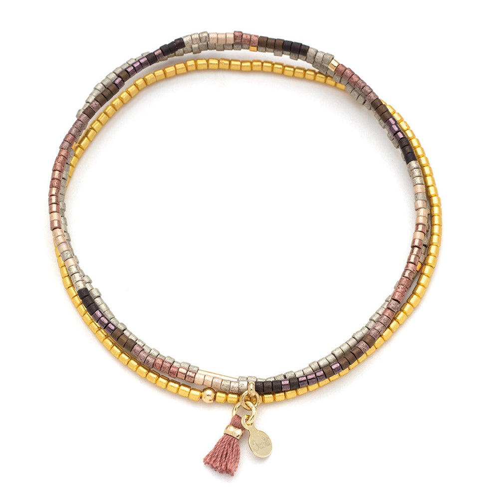 The Bronze Multi Gold Miyuki Bracelet Trio by Scout Jewelry features earthy-toned miniature square Miyuki Delica beads arranged in strands of gold, beige, black, and pink. This stretch cord bracelet is adorned with a small pink tassel and a tiny gold charm, making it perfect for stacking bracelet enthusiasts.