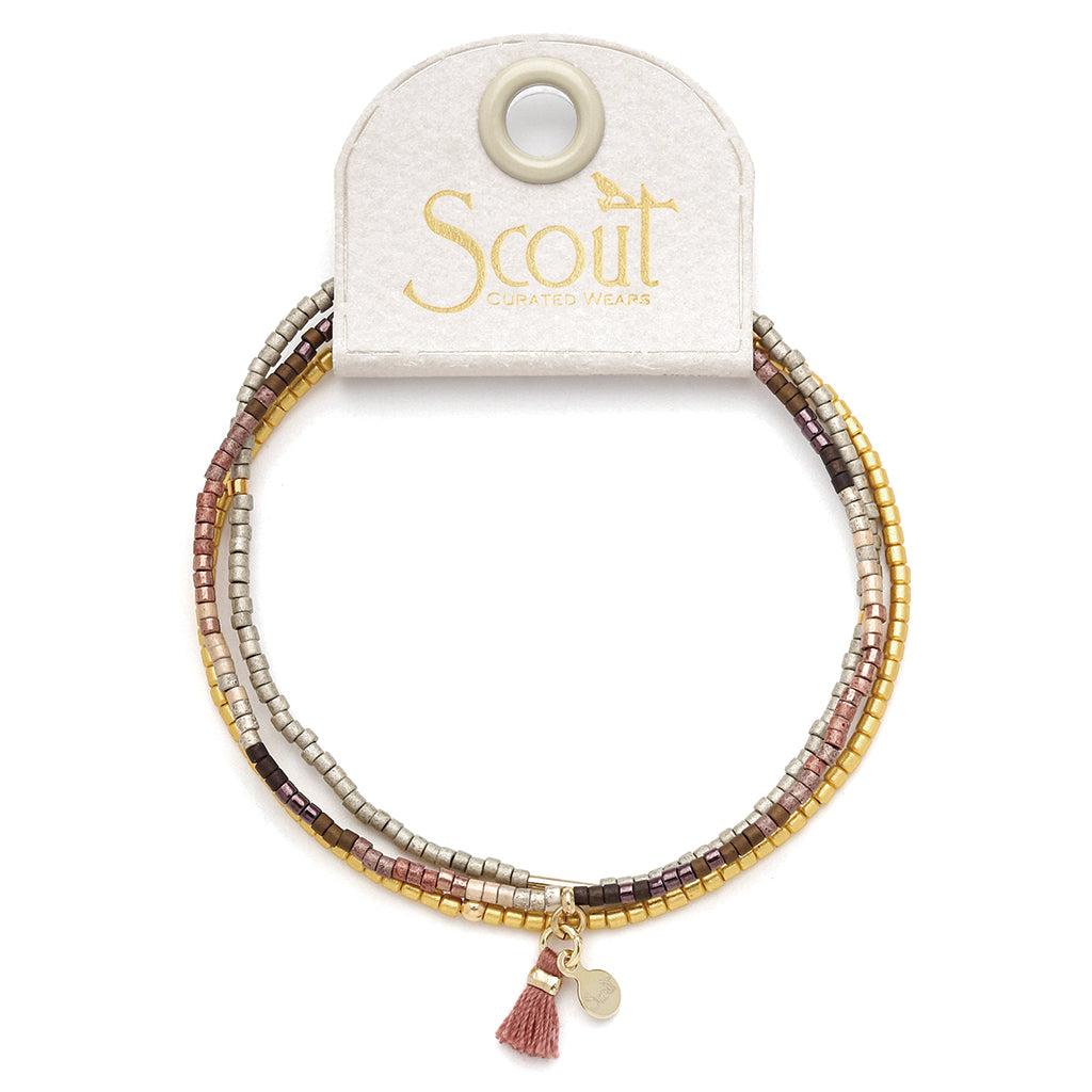 Introducing the Bronze Multi Gold Miyuki Bracelet Trio by Scout Jewelry, a multi-strand bracelet adorned with small square Miyuki Delica beads in shades of gold, silver, pink, and brown on a stretch cord. It features a tiny gold charm and a delicate pink tassel. The bracelet is elegantly displayed on a white tag bearing the Scout Jewelry logo.