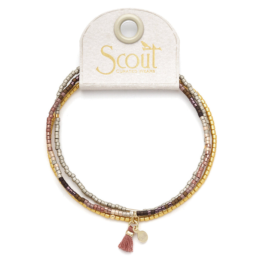 Introducing the Bronze Multi Gold Miyuki Bracelet Trio by Scout Jewelry, a multi-strand bracelet adorned with small square Miyuki Delica beads in shades of gold, silver, pink, and brown on a stretch cord. It features a tiny gold charm and a delicate pink tassel. The bracelet is elegantly displayed on a white tag bearing the Scout Jewelry logo.