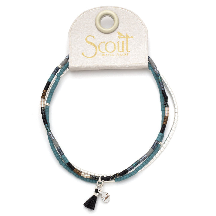 Introducing the Black Multi Silver Miyuki Bracelet Trio by Scout Jewelry, a set of stacking bracelets featuring Miyuki Delica beads in shades of blue, white, gray, and brown. This elegant trio is accented with a small Sterling Silver dipped charm and a black tassel, beautifully presented on a branded Scout Jewelry cardboard holder.