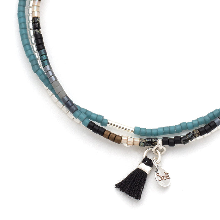 The Black Multi Silver Miyuki Bracelet Trio by Scout Jewelry is a multistrand stacking bracelet embellished with Miyuki Delica beads in hues of blue, black, gray, and silver. It includes a black tassel and a small Sterling Silver dipped charm engraved with the word "Scout.