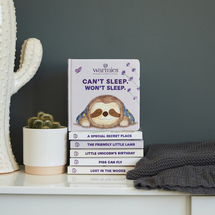 A cozy scene for sleepy readers features Warmies' plush sloth atop a children's book, "Can’t Sleep Won’t Sleep." A white cactus-shaped vase and a gray knitted blanket complete this Lavender Land against the dark wall.