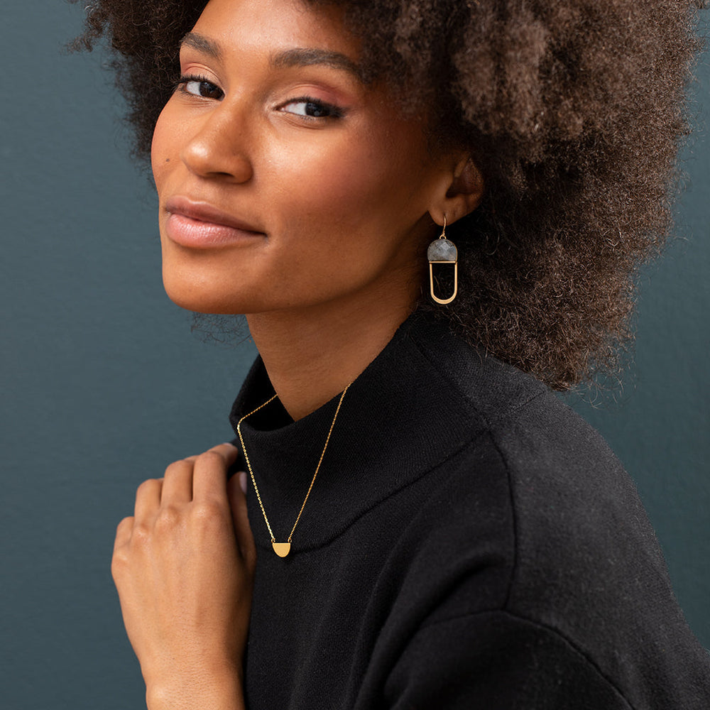 Against a dark teal background, a person with curly hair smiles softly while wearing a black top. Their ensemble features a gold necklace with a small pendant and is beautifully complemented by elegant, hypoallergenic sterling silver earrings from Scout Jewelry: the Modern Stone Earring Labradorite Gold, showcasing an exquisite stone and hoop design.