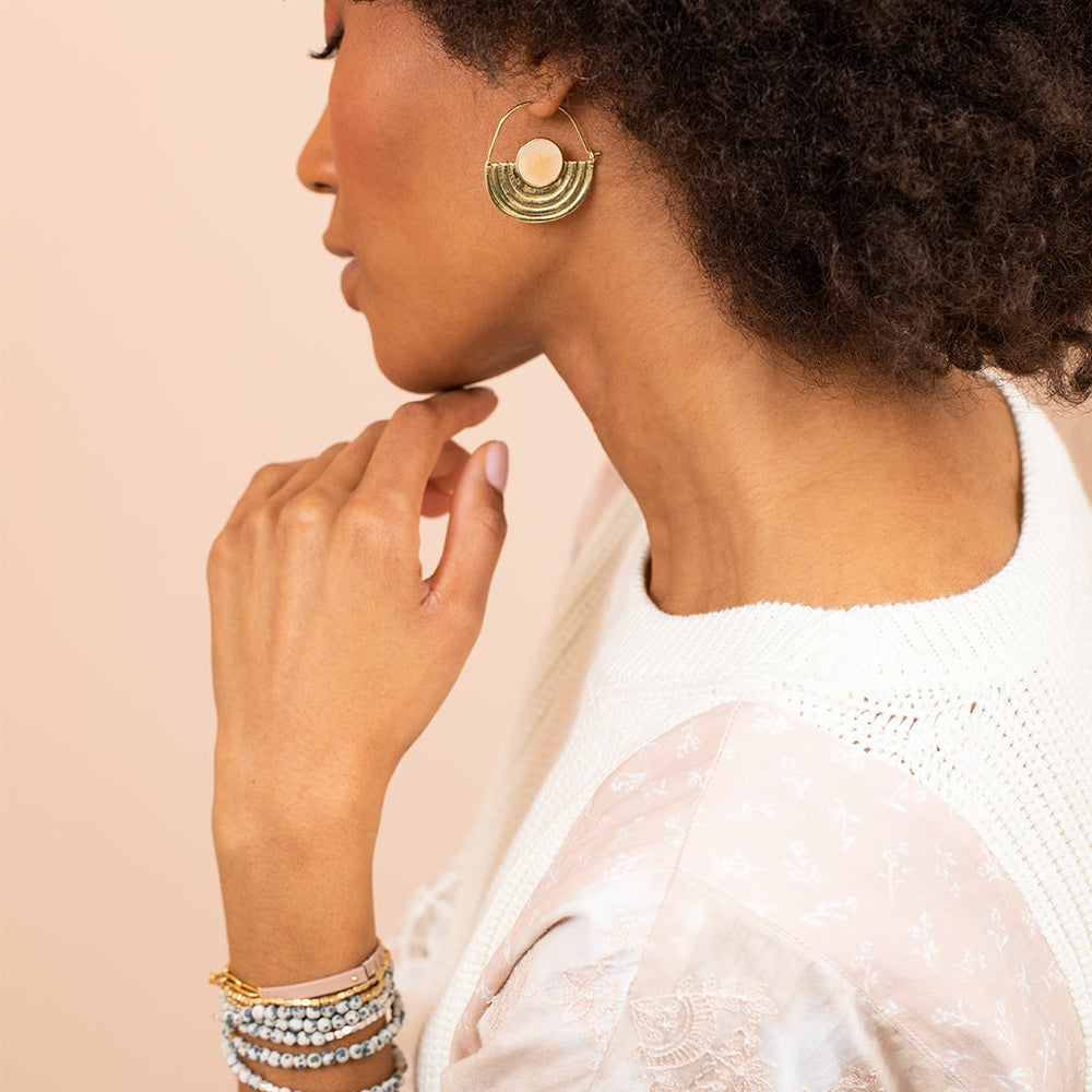 A woman with curly hair wearing a white and pink blouse and a knitted vest touches her chin. She has crescent-shaped earrings from Scout Jewelry, specifically the Stone Orbit Earring Sunstone Gold, which are hypoallergenic and dipped in 14K gold. On her wrist, she wears stacked bracelets. The background is a soft beige.