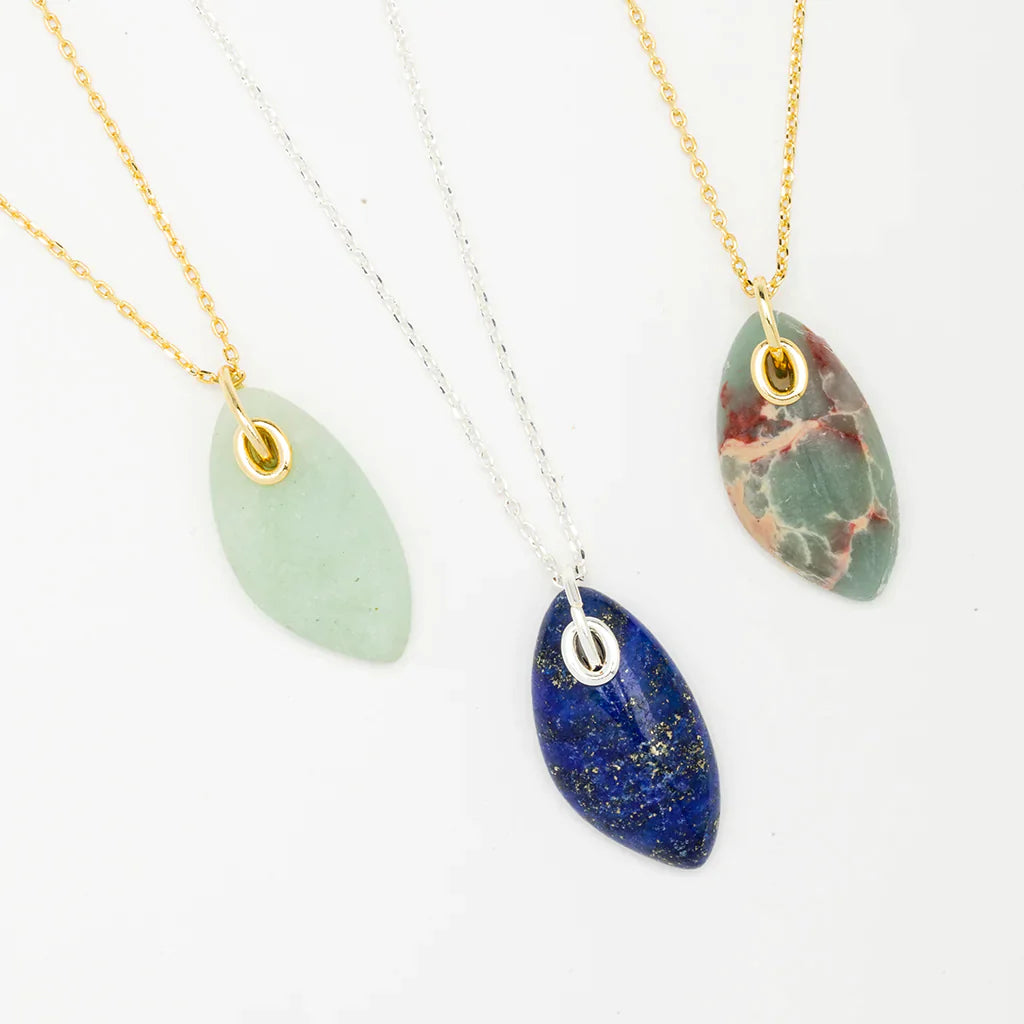 Three oval-shaped gemstone necklaces with thin adjustable chains displayed against a white background. The left necklace, featuring a light green stone on a 14K gold chain, the middle displaying the Lapis Silver Organic Stone Necklace by Scout Jewelry with a deep blue stone on a silver chain, and the right boasting a multi-colored stone on another 14K gold chain.