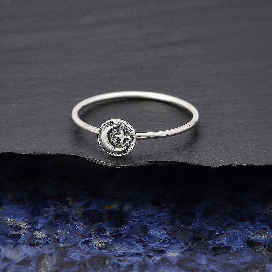The s7 Raised Moon and Star Stacking ring by Nina Designs is crafted from delicate sterling silver and showcases a small circular emblem with a crescent moon and star design, elegantly placed on a textured dark surface with subtle hints of blue.