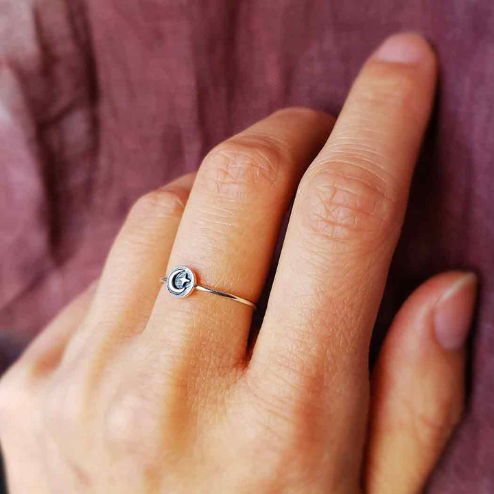 A hand adorned with the s8 Raised Moon and Star Stacking ring by Nina Designs, crafted in minimalist sterling silver featuring a small black and white letter 'G' set on a circular design. The backdrop is textured fabric in shades of purple.