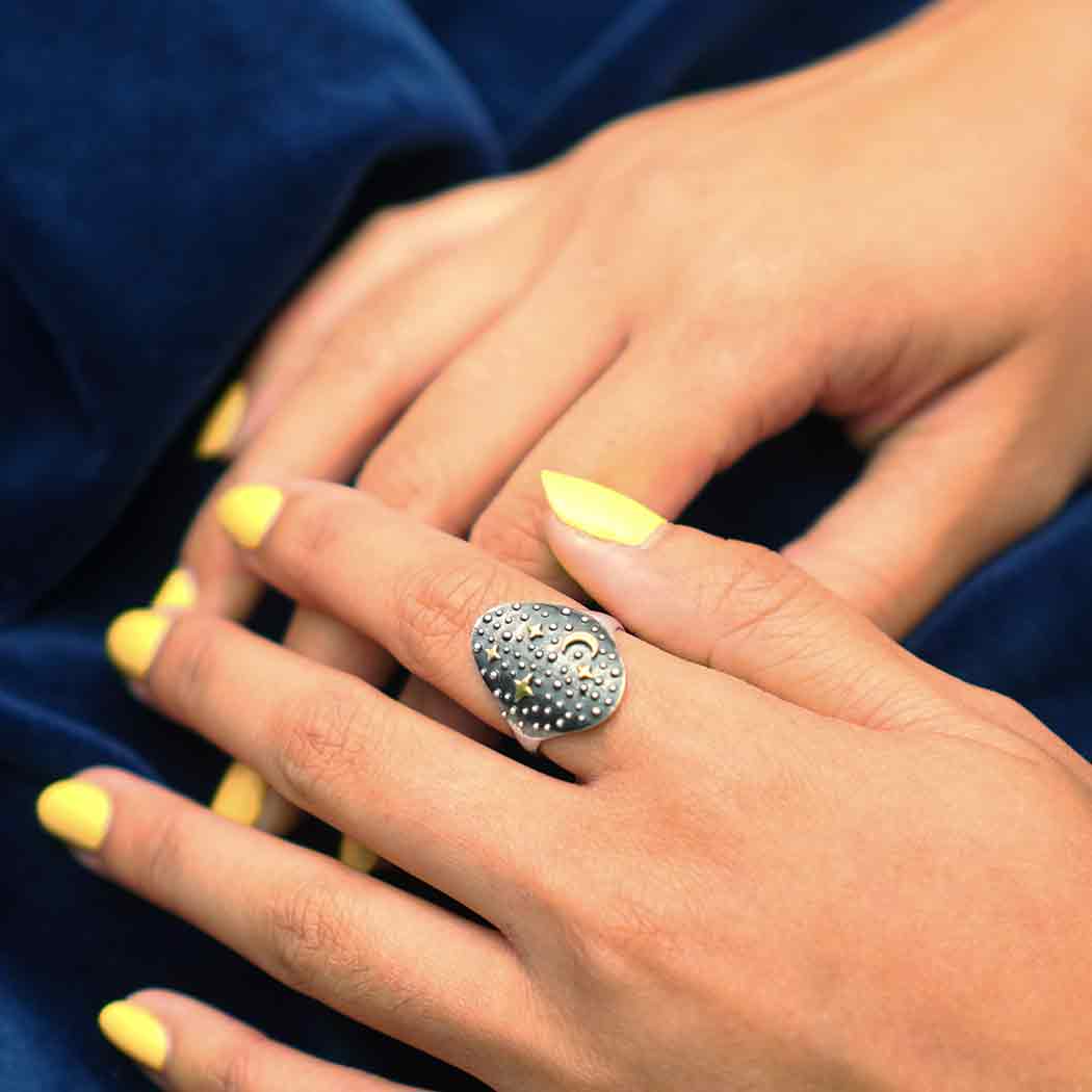 Close-up of hands with bright yellow nail polish, adorned with a large, textured sterling silver ring resembling the Nina Designs s6 Night Sky Ring with Moon and Stars, resting on a dark blue fabric background.