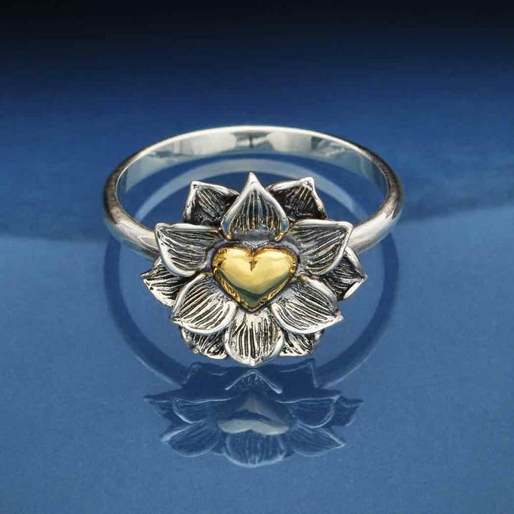 Set against a reflective blue surface, the s6 Lotus Ring with Heart by Nina Designs showcases intricate silver petals with delicate veining, gracefully surrounding a warm bronze heart at its center.