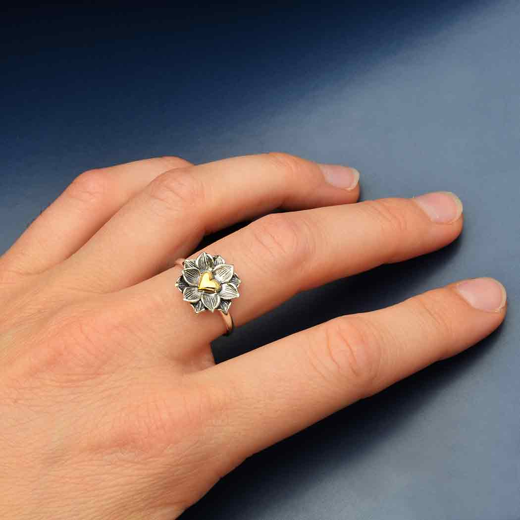 A hand adorned with the Nina Designs s5 Lotus Ring with Heart, featuring a silver flower shape with delicate veining and a bronze heart, set against a blue background.