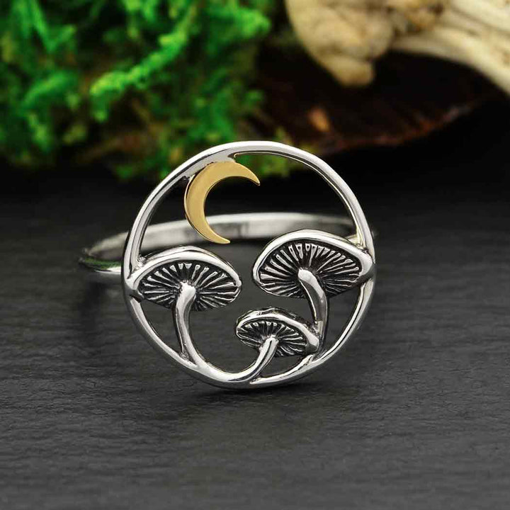 The Sz6 Mushroom Ring with Bronze Moon by Nina Designs features a round silver design adorned with two delicate mushrooms and a small bronze crescent moon. The background includes green moss and wood, capturing the essence of nature's healing properties and spirit medicine.