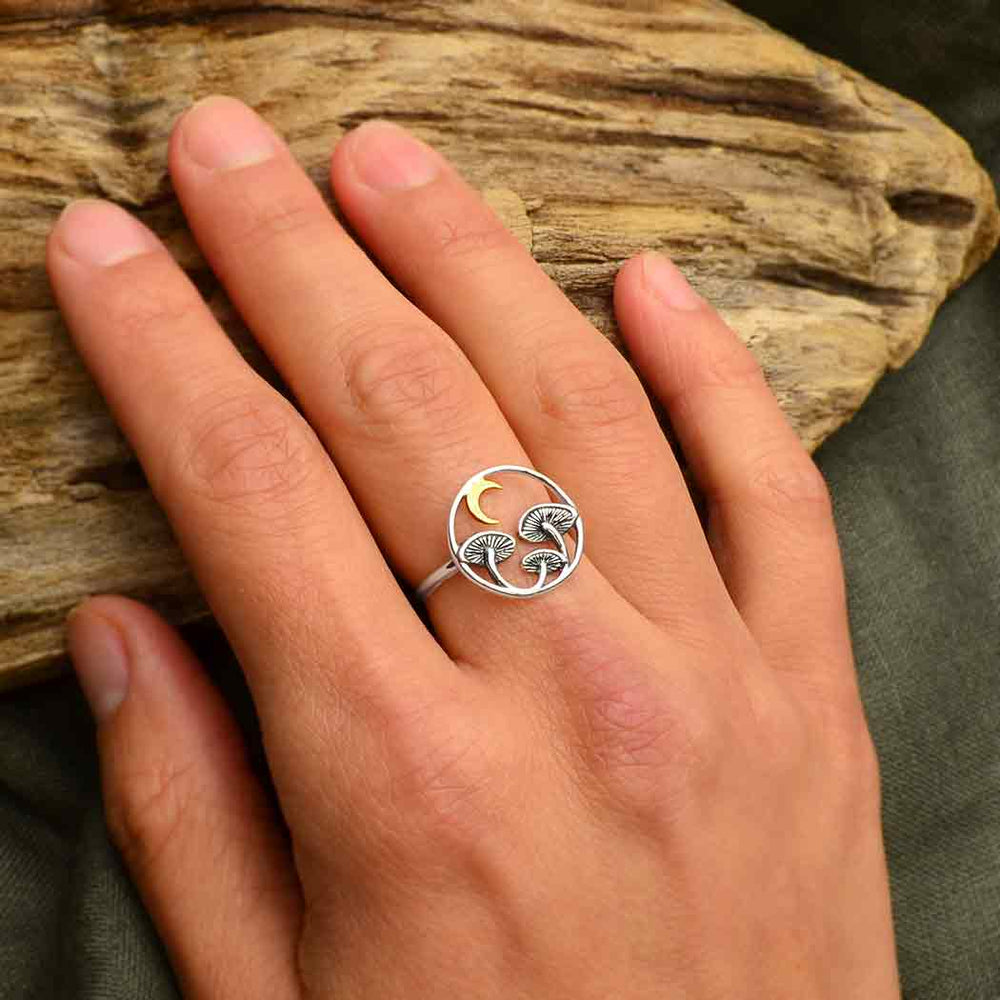 A hand adorned with Nina Designs' Sz5 Mushroom Ring with Bronze Moon, capturing the essence of spirit medicine with its three fungi and a delicate bronze crescent moon. The scene is set against driftwood resting on green fabric, evoking the healing properties of nature's gentle embrace.