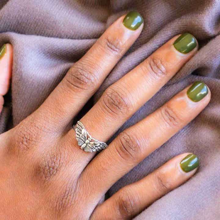 A hand with olive green nail polish rests on a soft gray fabric, adorned with the s8 Winged Butterfly Ring by Nina Designs, a sterling silver piece shaped like a butterfly on the middle finger.