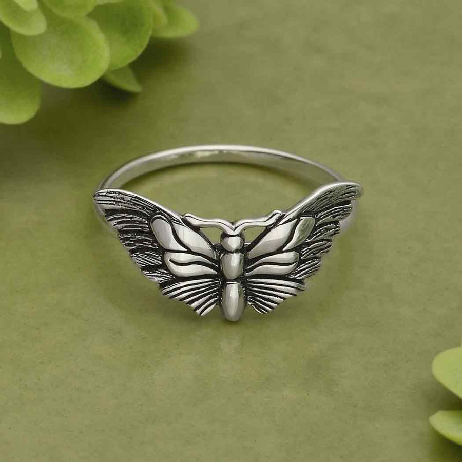 The s9 Winged Butterfly Ring by Nina Designs, crafted in sterling silver with an exquisitely detailed butterfly design, is showcased against a lush green background complemented by leaves in the corners. Its intricately designed wings exude elegance, enhancing intuition and psychic awareness with their captivating charm.