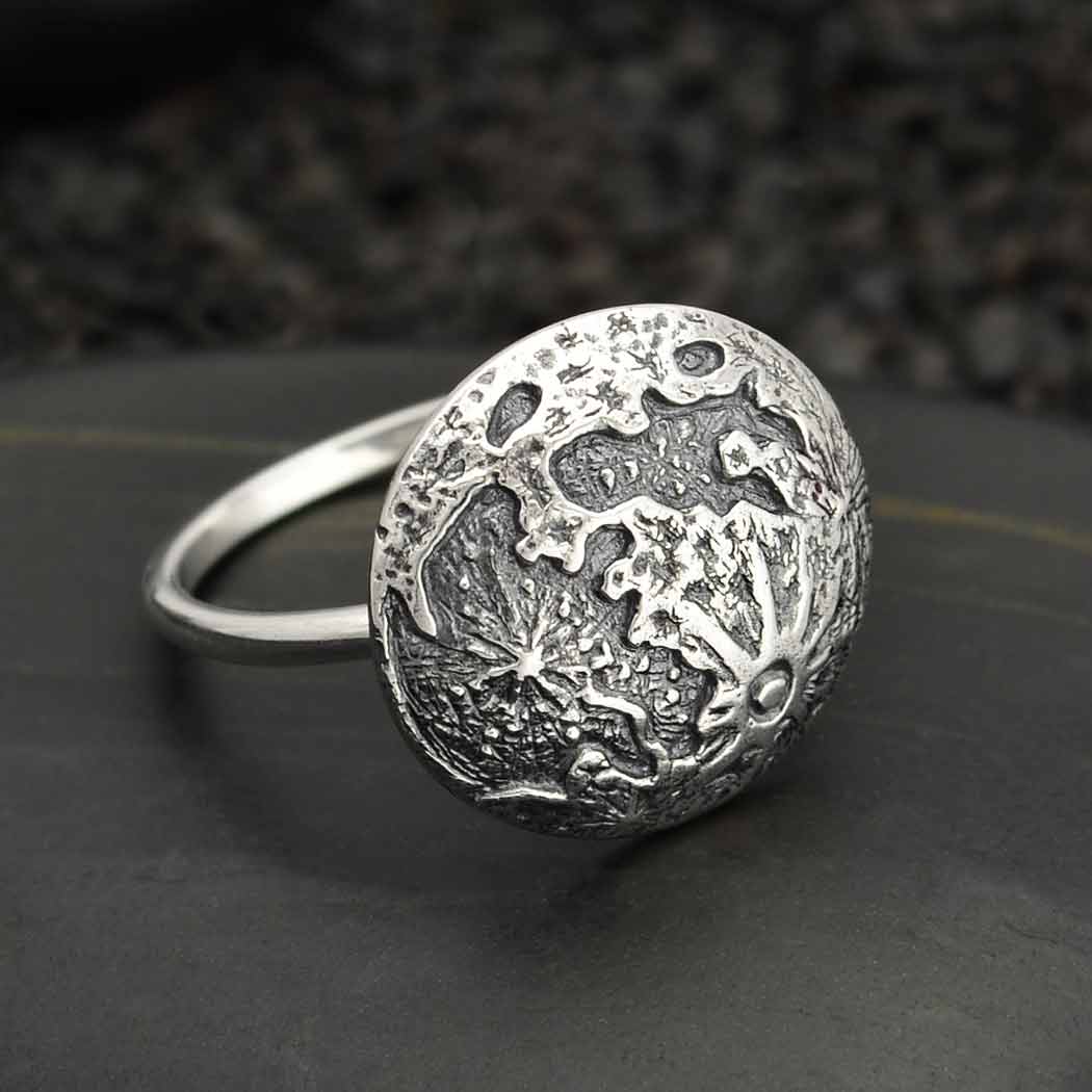 The Full Moon Silver Size 8 Ring by Nina Designs, crafted from sterling silver, features a detailed circular design reminiscent of textured craters or an abstract map. It is set against a dark textured surface, with intricate engravings and raised patterns that evoke the mystical beauty of a full moon, giving it a distinctive artisanal appearance.