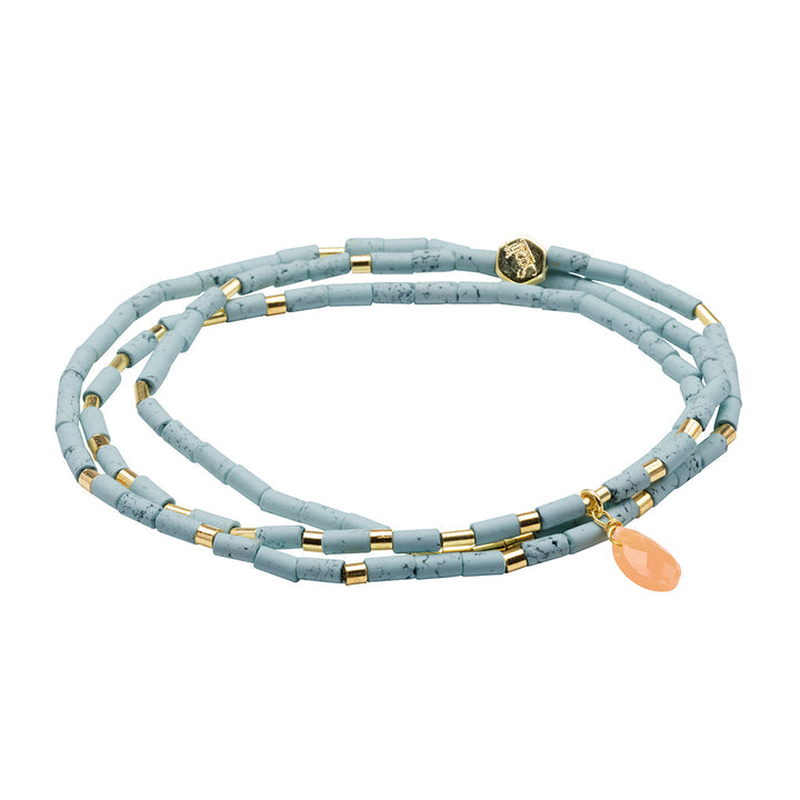 The Harmony Blue Howlite Gold Teardrop Wrap by Scout Jewelry is a striking blue beaded necklace, showcasing small cylindrical semi-precious stone beads with elegant gold accents and a peach faceted teardrop-shaped charm. The necklace is artistically coiled in a spiral on a white background.