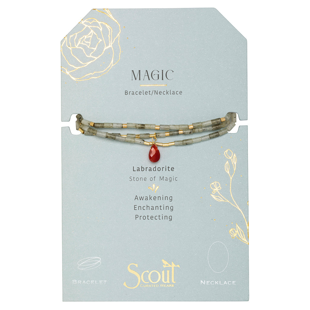 The packaging for the "Magic Labradorite Gold Teardrop Wrap" by Scout Jewelry is gray and accompanies a bracelet or necklace adorned with labradorite semi-precious stone beads and a red charm. It features text that highlights "Labradorite: Stone of Magic" along with words like "Awakening, Enchanting, Protecting." The package is beautifully decorated with gold floral designs and plated brass accents.