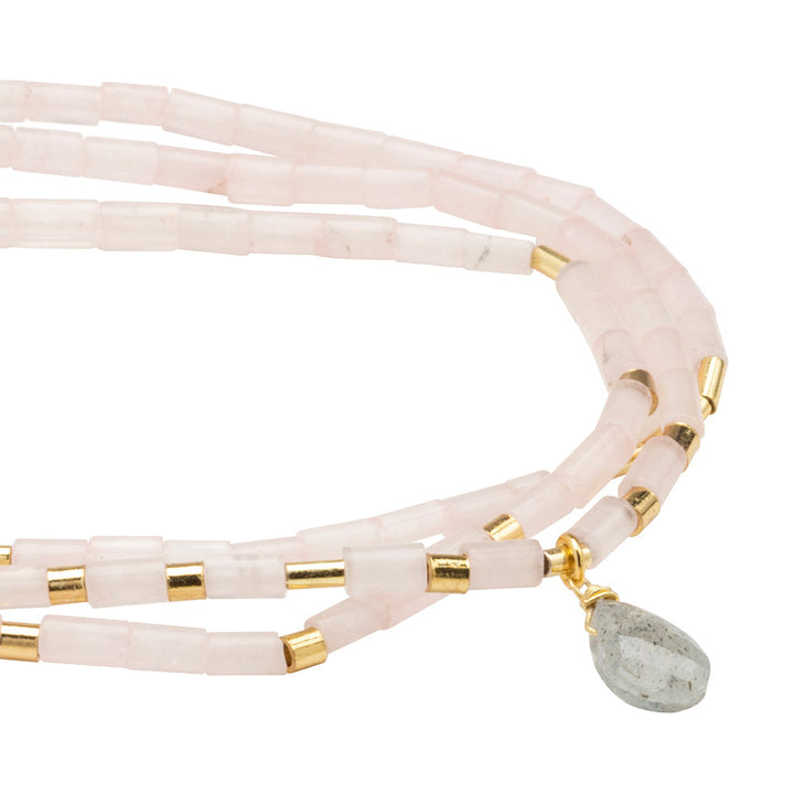 The Love Rose Quartz Gold Teardrop Wrap by Scout Jewelry is a multi-strand bracelet with pale pink cylindrical rose quartz beads, small gold accents, and a dangling teardrop-shaped semi-precious stone. It's simple and elegant.