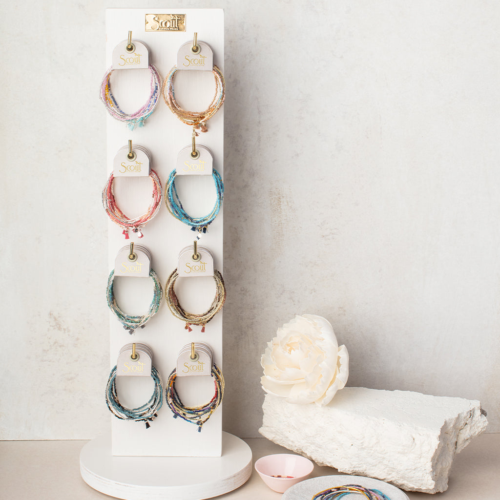 A white vertical display stand holds nine sets of the Turquoise Multi Silver Miyuki Bracelet Trio from Scout Jewelry, showcasing colorful beaded stacking bracelets with intricate Miyuki Delica beads on stretch cords. The stand is complemented by a textured white stone, an artificial white peony to the right, and a small pink dish in the corner.