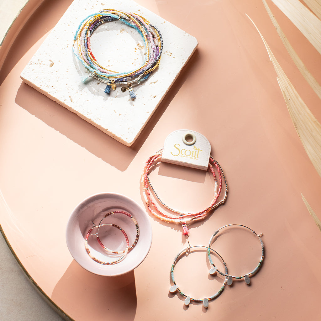 A pink tray showcases a collection of jewelry, including the Cobalt Multi Gold Miyuki Bracelet Trio, crafted with multicolored beads. The display also features a pair of large 14K gold hoop earrings and a delicate set with dangling charms. A tag displaying the brand name "Scout Jewelry" is visible as bright sunlight highlights these treasures.